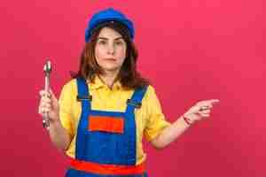 Foto gratuita uniforme di costruzione d'uso della donna turbata del costruttore e chiave di tenuta del casco di sicurezza a disposizione che indicano dito il lato che controlla parete rosa isolata