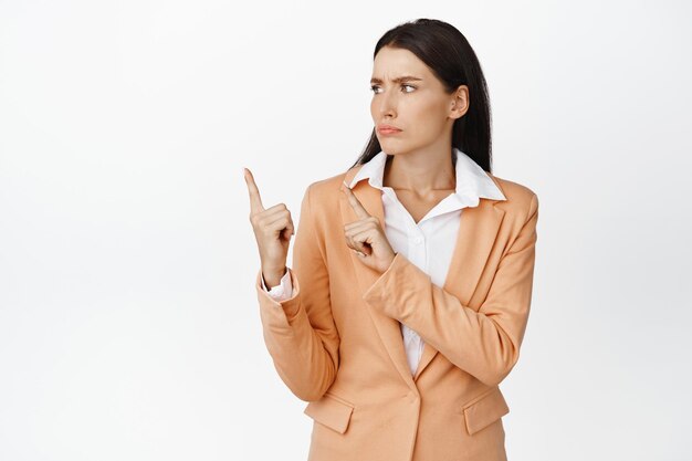 Upset businesswoman in suit pointing fingers at upper left corner and looking disappointed standing against white background