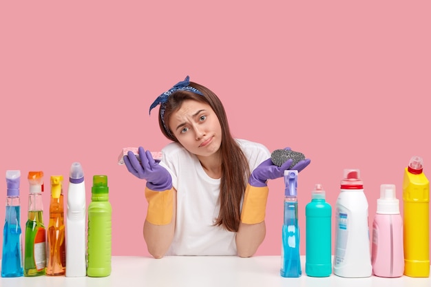 Upset Caucasian woman tilts head, purses lips, holds sponge, surrounded with cleanser and other chemical supplies