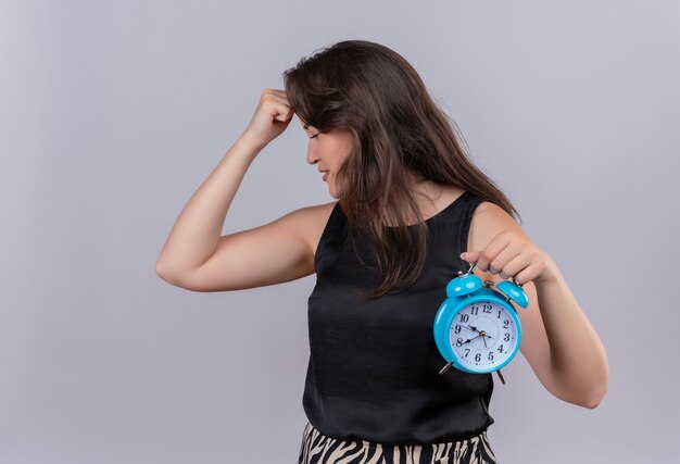 Upset caucasian young girl wearing black undershirt holding a alarm clock and put her hand on forehead on white background