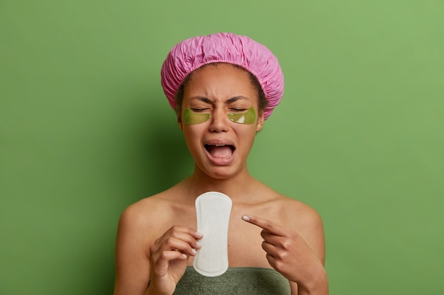 Бесплатное фото Расстроенный крик от отчаяния указывает на гигиеническую салфетку, носит банную шапку, накладывает коллагеновые пластыри под глаза, позирует на зеленой стене, чувствует боль во время менструации