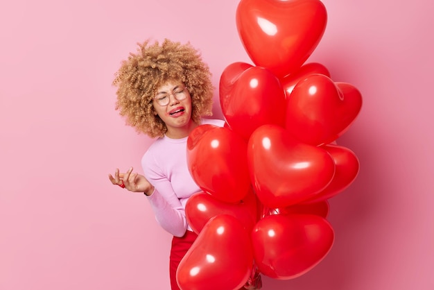 Free photo upset crying woman has spoiled makeup curly hair feels dejected holds bunch of heart balloons keeps hand raised expresses negative emotions isolated over pink background festive event concept