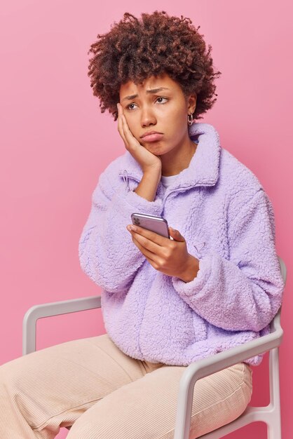 Upset curly haired woman sits on chair uses mobile phone received bad sms feels very unhappy wears fur jacket and trousers isolated over pink wall