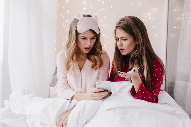 Upset curly woman in eyemask looking at phone screen. Interested long-haired girl sitting under blanket with smartphone.