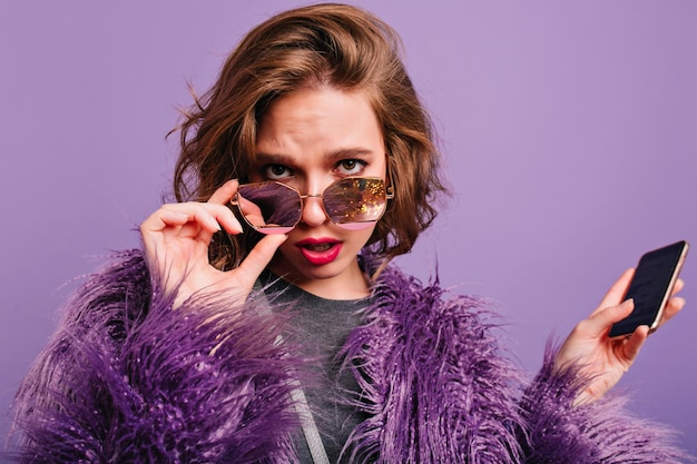 紫色の背景にサングラスを通して見ているスマートフォンを手に動揺した巻き毛の女性。写真撮影でポーズをとって明るいメイクでファッショナブルな冬の服装で魅力的な女の子。