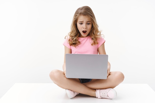 Upset cute blond little kid getting sad and disappointed checking school account receive terrible mark sit crossed legs laptop open mouth gasping look computer screen shocked white background