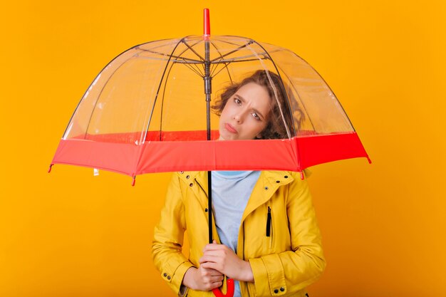 Upset dark-haired girl posing under umbrella . Portrait of sad caucasian lady in raincoat holding parasol on bright wall.
