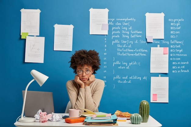 Upset dark skinned worker holds chin, sits at desk near laptop computer analyzes results feels tired of working.