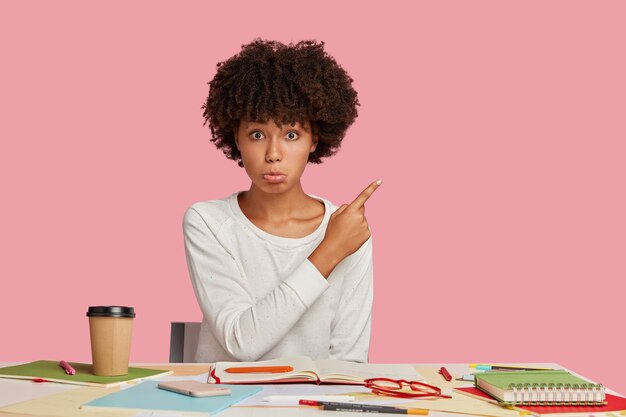 Upset designer purses lips in discontent, sits at desktop, drinks beverage from disposable cup, points at copy space