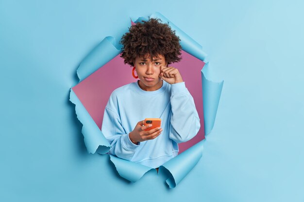 Upset disappointed curly woman wipes tears unhappy as boyfriend doesnt call her holds modern mobile phone in hands expresses negative emotions breaks through blue paper wall
