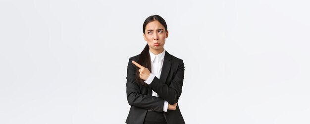 Upset and disappointed female entrepreneur broker in suit grimacing pouting displeased and sad as looking and pointing upper left corner uneasy standing white background