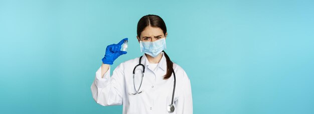 Upset and disappointed woman doctor in face medical mask showing vaccine with sad dislike face stand