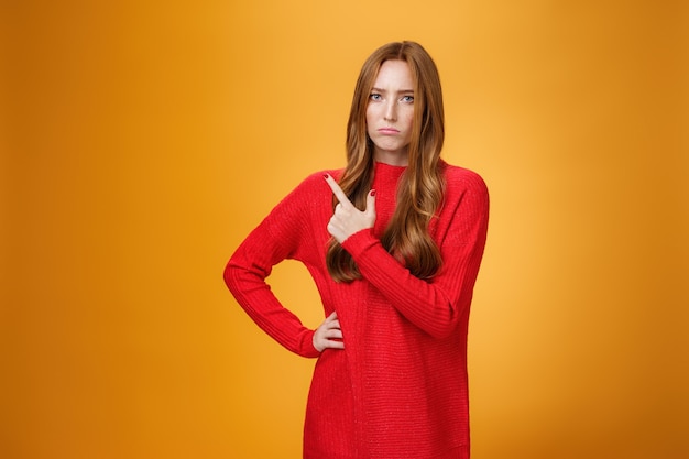 Upset and disappointed young ginger girl in red sweater pointing at upper left corner or behind frowning looking gloomy and angry, standing jealous and moody against orange background