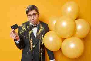 Free photo upset discontent man holds bank ccard has no money for celebration looks sadly purses lips wears formal clothes hods bunch of inflated balloons isolated over yellow background holidays concept