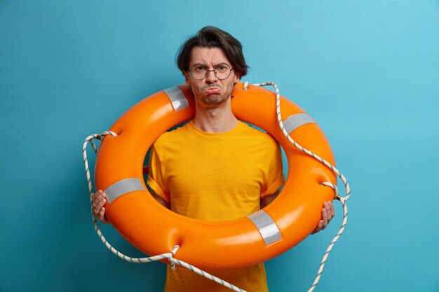 Расстроенный недовольный взрослый мужчина несет кольцо-спасатель, носит прозрачные очки и оранжевую футболку