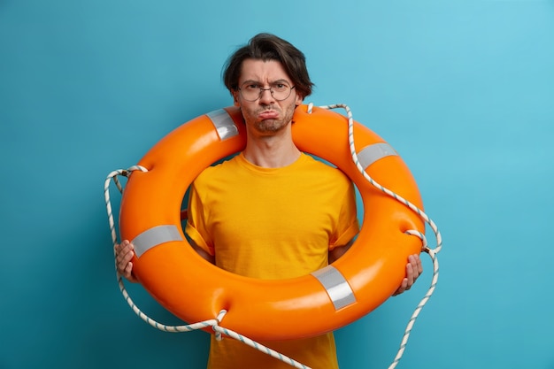 upset displeased adult man carries ring lifesaver, wears transparent glasses and orange t shirt