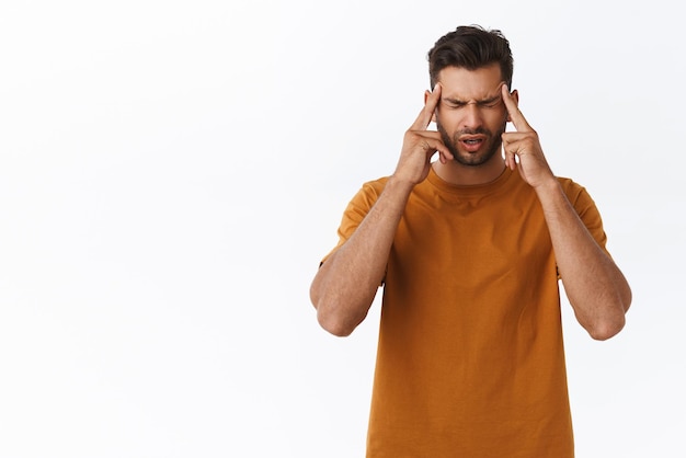 Free photo upset and distressed tensed young bearded guy suffering terrible migraine massaging temples with index fingers close eyes and grimacing from pain painful headache standing white background