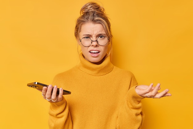 Sconvolto disturbato insoddisfatto giovane donna con i capelli biondi raccolti in chignon solleva palmo alza le spalle indossa occhiali rotondi maglione casual tiene cellulare non riesce a capire come inviare un messaggio