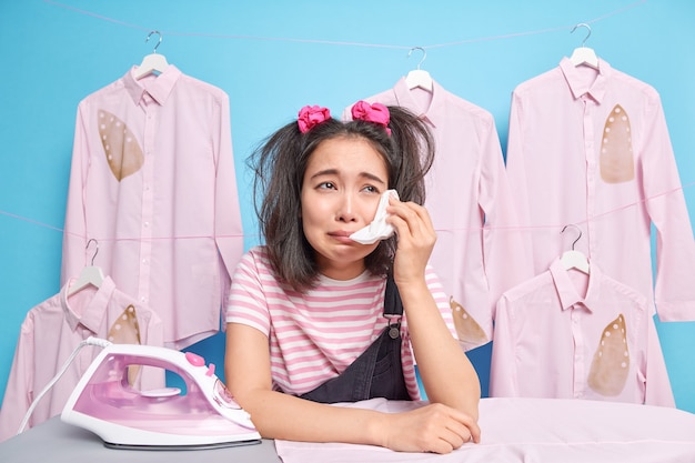 Upset doleful Asian teenage girl has two pony tails wipes tears with handkerchief leans at ironing board poses near ironed clothes on hangers finds out bad new feels tired of daily domestic routines