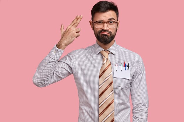 Upset European man with thick bristle makes suicide gesture, shoots himeslf in temple, has displeased facial expression, wears formal outfit