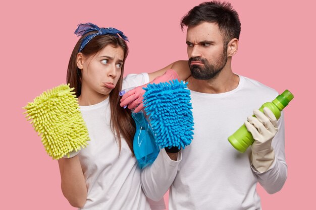 Upset European woman and man frown faces while look at each other, hold spray and bottle of detergent, colourful rags