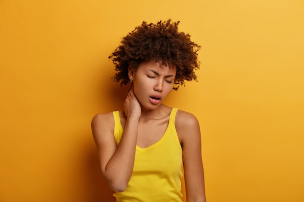 Upset fatigue curly young woman touches neck, suffers from painful feelings after sitting long time at computer, frowns face, wears yellow casual clothes, has problems with spine, poses indoor