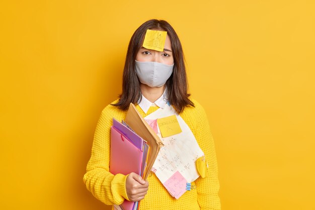 検疫中に遠方で働く動揺した女子学生は、保護マスクを着用し、グラフィックが描かれた粘着性のメモを着用して、プロジェクトの作業が悲しそうに見えるように準備します。