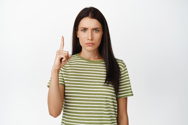 Расстроенная хмурая девушка, указывающая пальцем вверх, выглядит ревнивой или разочарованной, стоя в футболке на белом фоне