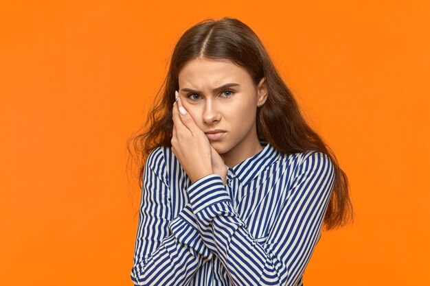 Upset frowning young European woman in striped shirt holding hand on her cheek because of toothache.