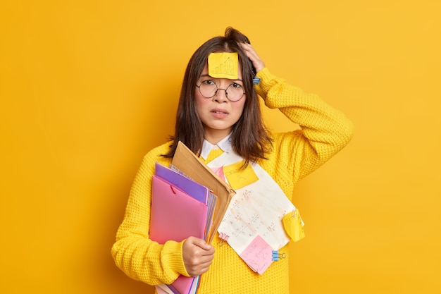 Upset frustrated office worker puzzled to have much work has sticker on forehead tries to do everything in time holds folders with papers wears big round spectacles.