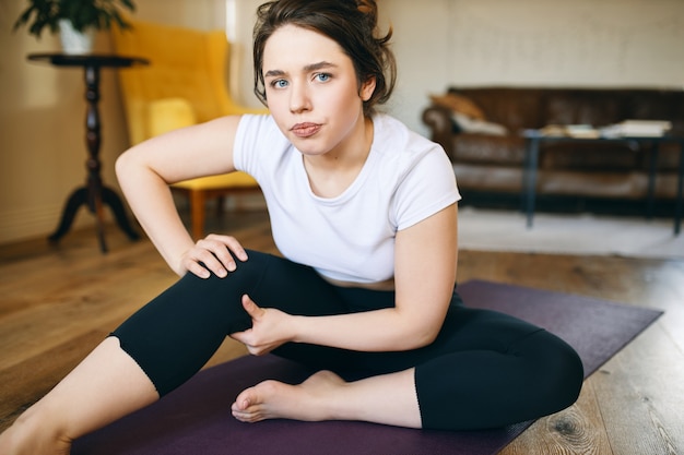 운동복을 입은 젊은 여성이 운동을 잘못했기 때문에 요가 수업 중에 무릎에 불편 함, 긴장 및 통증을 느끼는 화가 좌절했습니다.