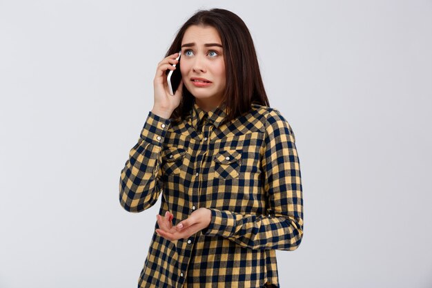 La giovane bella ragazza divertente turbata si è vestita in camicia di plaid che parla sul telefono, esaminando il lato sopra la parete bianca.