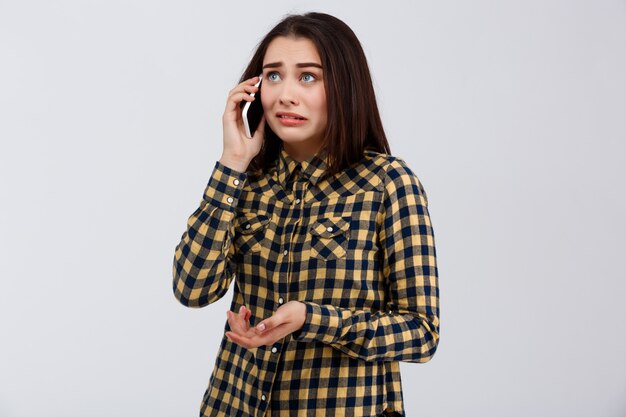 Upset funny young beautiful girl dressed in plaid shirt speaking on phone, looking at side over white wall.