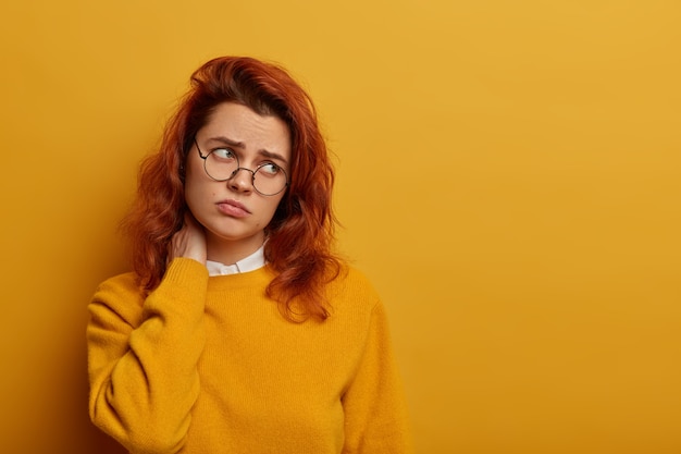 Upset ginger woman tilts head aside, suffers from neck pain, looks with dissatisfaction aside, wears yellow jumper, roung spectacles, leads sedentary lifestyle, needs massage