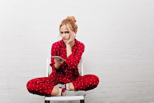 Upset girl in cute socks sitting on chair and waiting for call. Indoor portrait of blonde sad young woman posing with legs crossed and holding smartphone.