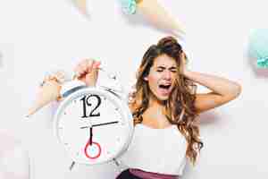 Free photo upset girl scream with eyes closed touching head in panic and holding big white clock. portrait of distressed young woman in white tank-top