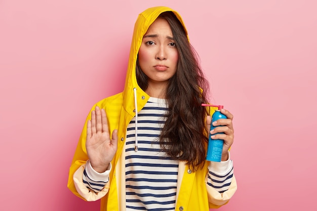 Upset gloomy Asian woman makes refusal gesture, says no, holds medical spray for avoiding illness, wears waterproof yellow raincoat with hood, striped jumper