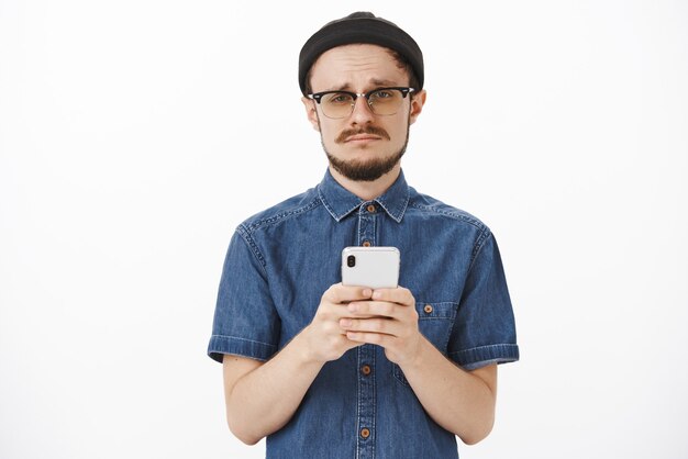 upset gloomy cute bearded guy in black beanie and glasses making sad face and frowning holding smartphone expressing jealousy or regret missing chance to buy tickets online