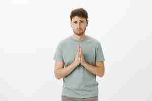 Free photo upset gloomy cute guy with bristle in t-shirt and earrings, holding hands in pray and making sad smile while asking for favour, being desperate