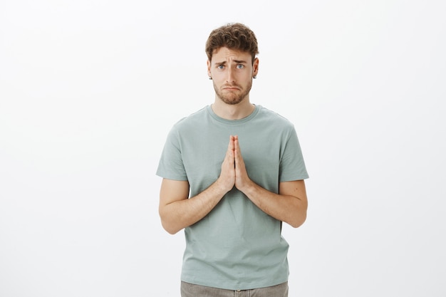 Upset gloomy cute guy with bristle in t-shirt and earrings, holding hands in pray and making sad smile while asking for favour, being desperate