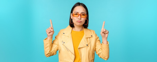 Upset gloomy korean woman in sunglasses points fingers up looks disappointed while shows banner on top stands over blue background