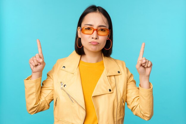 Upset gloomy korean woman in sunglasses points fingers up looks disappointed while shows banner on top stands over blue background