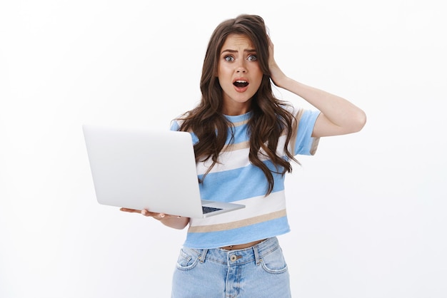 Free photo upset gloomy troubled cute woman hold laptop grab head, gasping worried, drop jaw and panicking have problem with computer