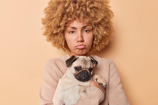 Sconvolto cupo giovane donna posa con pug dog sulle mani ha cattivo umore come il suo animale domestico è malato andando a visitare le labbra di borse veterinarie si sente deluso vestito casualmente isolato su sfondo beige.