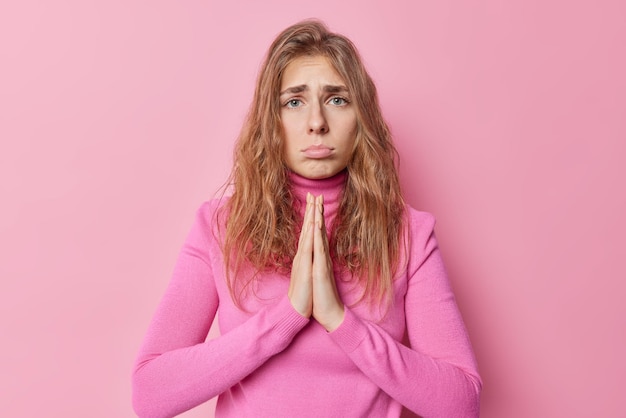 Free photo upset good looking young woman with needy face begsfor something stands in pray pose looks sadly purses lips wears casual turtleneck isolated over pink background. please give me one more chance