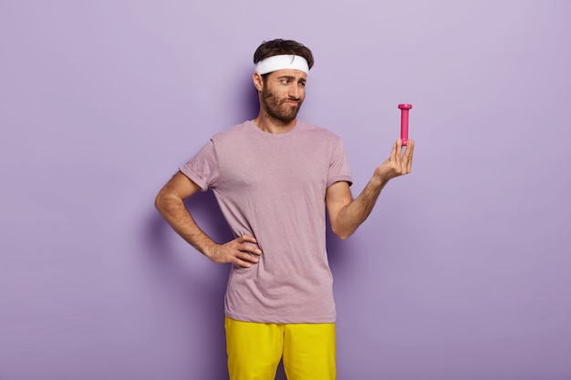 Upset guy looks with apathy at small dumbbell in his hand, thinks its too light, wears headband