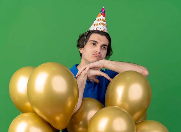 Расстроенный красивый кавказский мужчина в кепке на день рождения стоит с гелиевыми шарами