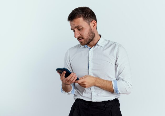 Upset handsome man holds and looks at phone isolated on white wall