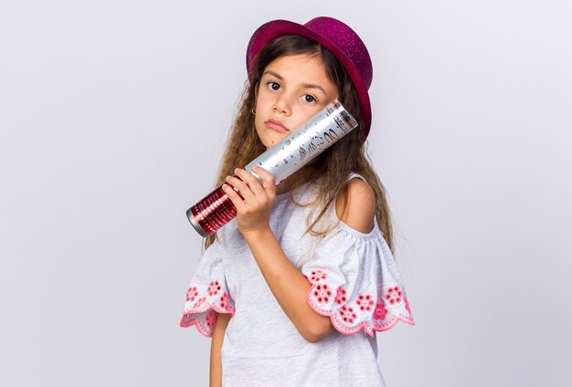 upset little caucasian girl with purple party hat holding confetti cannon isolated on white wall with copy space