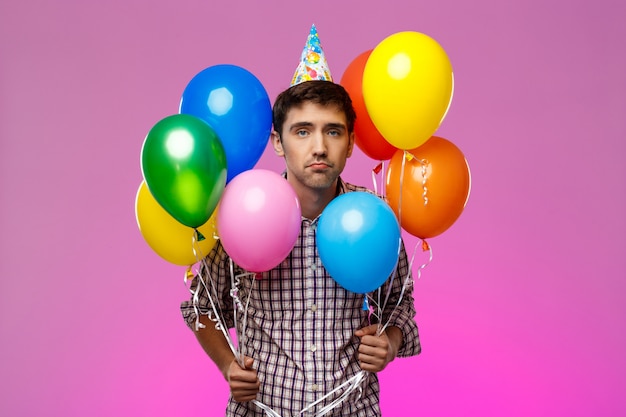 Upset man celebrating birthday, holding colorful baloons over purple wall.